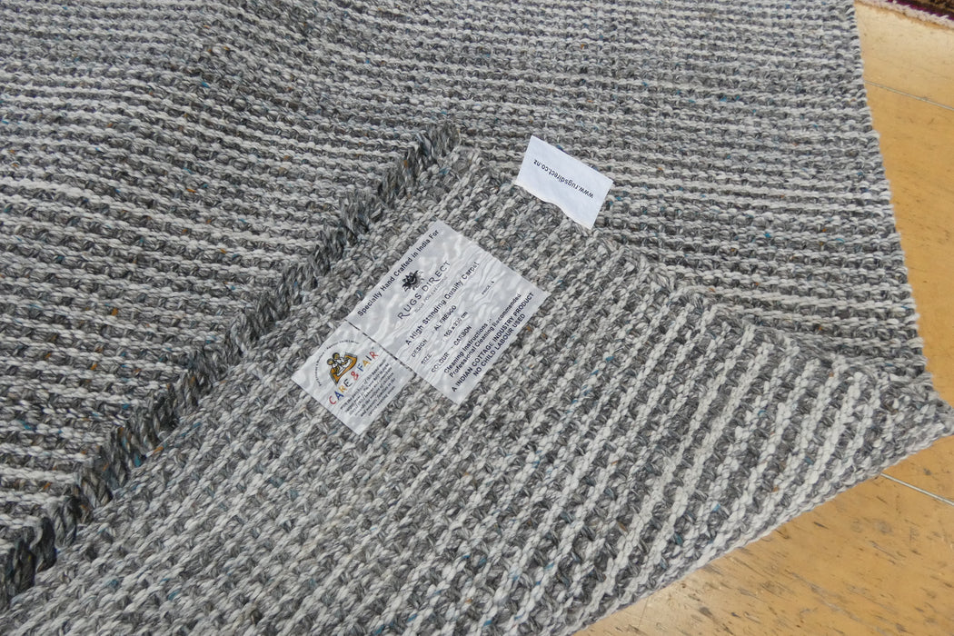 A close-up of a gray and white textured woven rug or carpet, folded over to show the underside with two white labels, placed on a light wood floor.