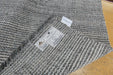 A close-up of a gray and white textured woven rug or carpet, folded over to show the underside with two white labels, placed on a light wood floor.