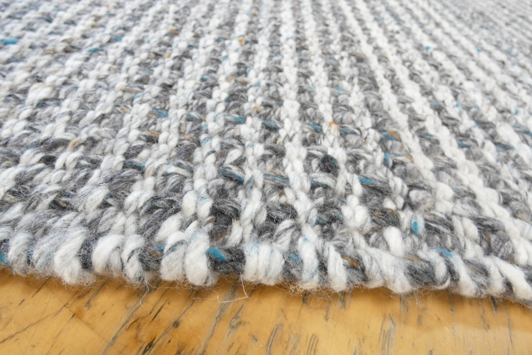 Close-up of a woven rug or textile with a chunky variegated texture in white, gray, black, and small blue flecks, resting on a light wood surface.