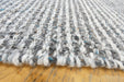 Close-up of a woven rug or textile with a chunky variegated texture in white, gray, black, and small blue flecks, resting on a light wood surface.