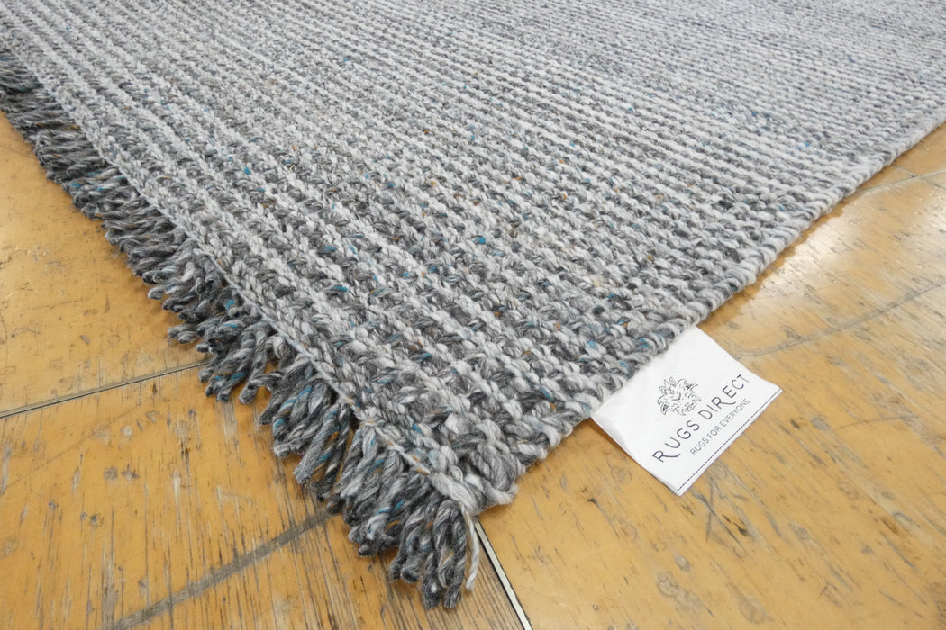 A close-up of a gray and white textured woven rug with fringed edges, laid on a light wood floor. A white fabric tag with the 'RUGS DIRECT' brand logo is visible near the corner.