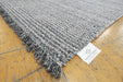 A close-up of a gray and white textured woven rug with fringed edges, laid on a light wood floor. A white fabric tag with the 'RUGS DIRECT' brand logo is visible near the corner.