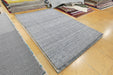A rectangular gray woven area rug with a textured, ribbed surface, displayed on a light-colored tiled floor in a showroom with other rugs stacked in the background.