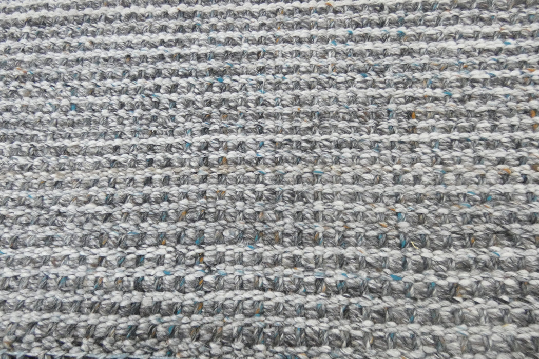 A close-up of a woven rug or mat with a textured, chunky pattern in heathered gray, white, and black, with subtle hints of blue and brown threads.