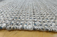 Close-up of a woven rug or textile with a chunky braided texture in off-white, gray, and small blue accents, shown resting on a light wood surface.
