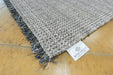 A close-up of a woven area rug with dark gray fringe, laid on a light wood floor. A white fabric tag with the 'RUGS DIRECT' brand name is visible near the corner.