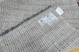 Close-up of a woven area rug with a textured pattern of beige, gray, and white threads, showing a white product label and a small white tag.