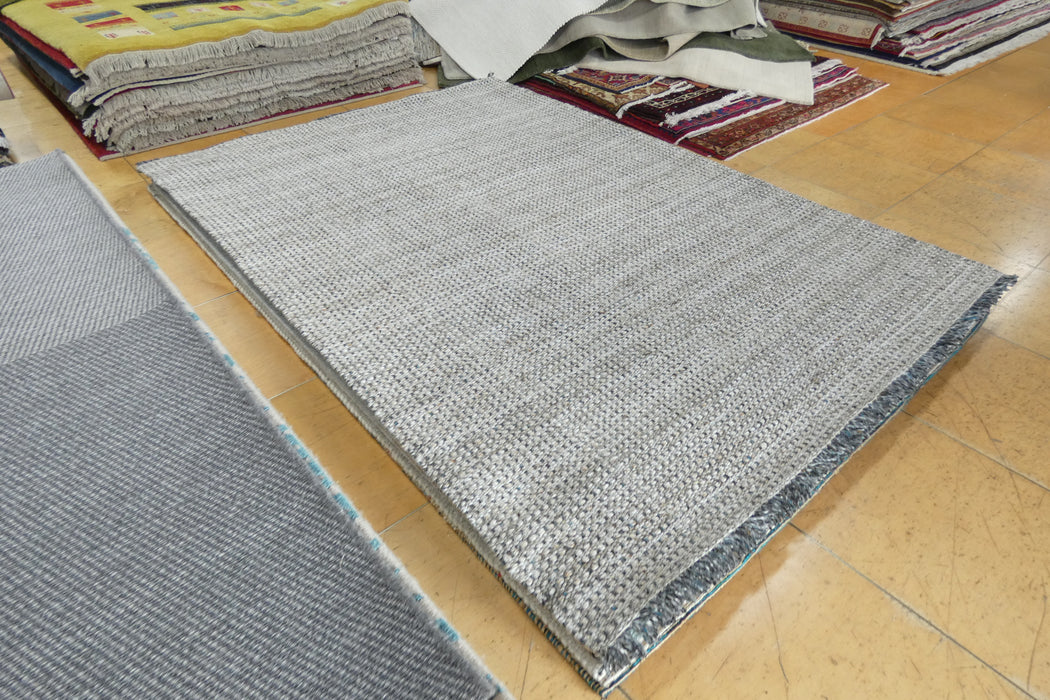 A rectangular light gray woven area rug laid out on a polished wood floor, with other rugs stacked in the background.