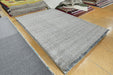 A rectangular light gray woven area rug laid out on a polished wood floor, with other rugs stacked in the background.