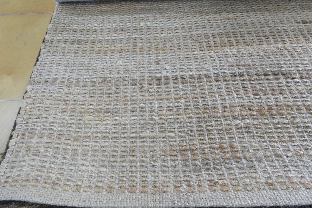 A close-up of a woven area rug with a textured basketweave pattern in beige, cream, and light brown tones, shown laid out on a floor.