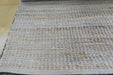 A close-up of a woven area rug with a textured basketweave pattern in beige, cream, and light brown tones, shown laid out on a floor.