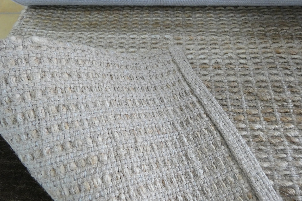 A close-up of a woven area rug with a textured, basketweave pattern in cream and light tan tones.