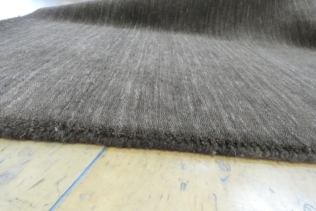 A close-up of a dark brown textured rug with a fringed edge, laid on a light-colored wooden floor.