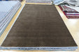 A large rectangular brown rug laid flat on a light tiled floor, with stacks of other rugs visible along the left and right edges.