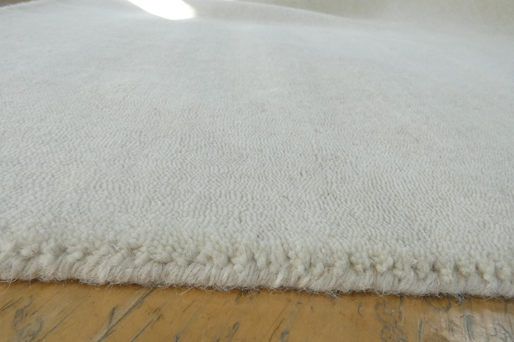 A close-up of a plush, off-white or cream-colored rug with a textured loop pile and a fringed edge, resting on a light-colored wooden floor.
