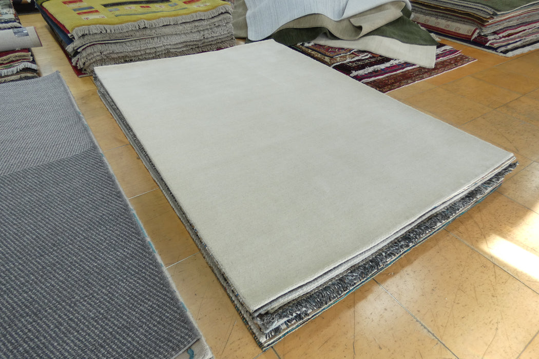 A large rectangular light beige rug or carpet sample displayed on a wooden floor, with other rugs stacked around it in a showroom.