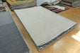 A large rectangular light beige rug or carpet sample displayed on a wooden floor, with other rugs stacked around it in a showroom.