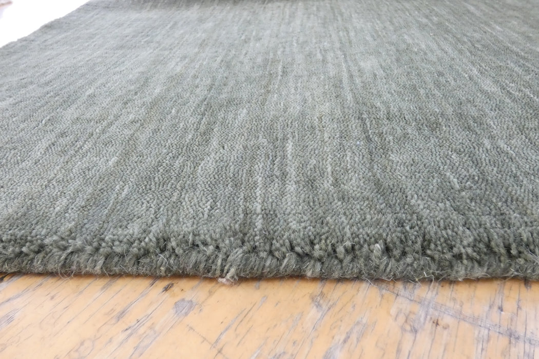 A close-up of a textured gray woven rug with a frayed edge, laid on a light-colored wooden floor.