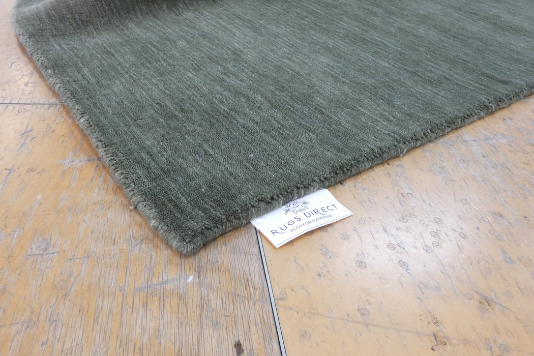 A close-up of a dark olive green textured rug corner with a visible 'RUGS DIRECT' tag, placed on a worn wooden floor.