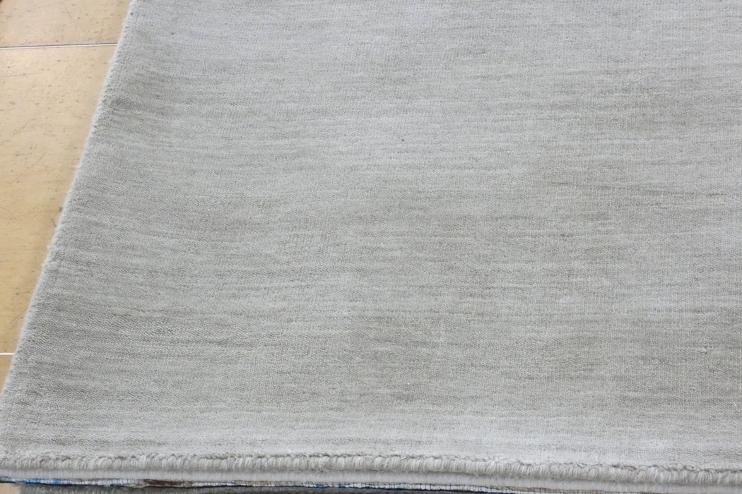 A close-up of a light gray textured rug with a visible fringed edge, laid on a light wood floor.