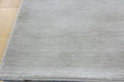 A close-up of a light gray textured rug with a visible fringed edge, laid on a light wood floor.