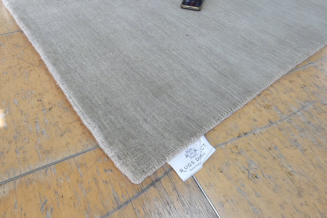 A close-up of a light gray rug laid on a worn wooden floor, with a small white fabric tag reading RUGS ROOIS visible near the corner.
