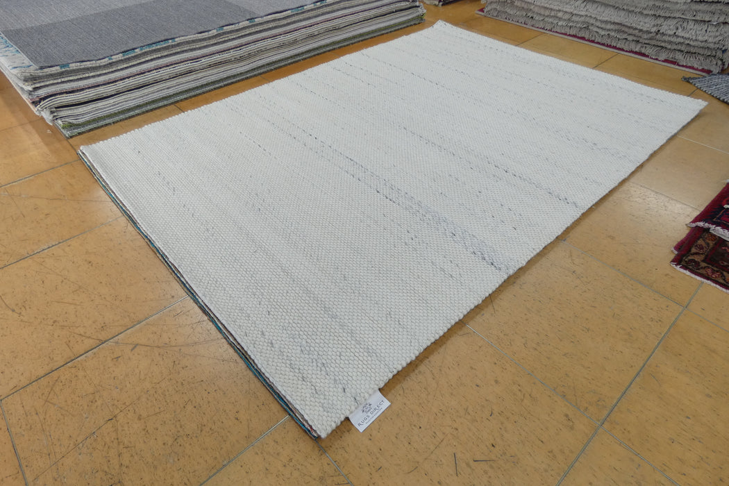 A large off-white woven area rug with subtle horizontal textured stripes, laid out on a tan tiled floor with other rugs stacked in the background.