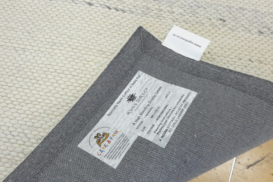 Close-up of the underside of a gray woven rug, showing a sewn-in label for 'Rug Company' and a small white size tag, laid on a textured cream-colored rug.