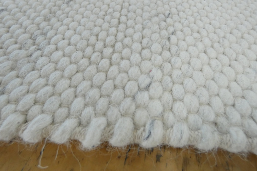 A close-up of a white textured woven rug with a chunky, looped tufted pattern and frayed edges, laid flat on a wooden surface.