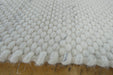 A close-up of a white textured woven rug with a chunky, looped tufted pattern and frayed edges, laid flat on a wooden surface.