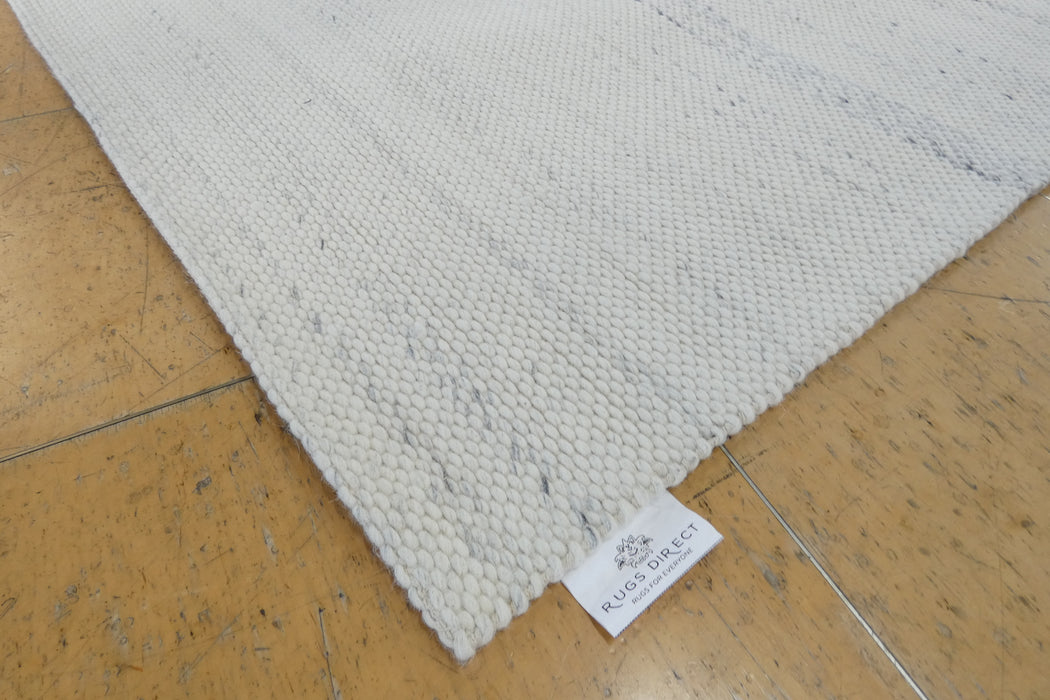 A close-up of a woven area rug corner on a polished wood floor, showing a white woven label that reads 'RUGS DIRECT'.