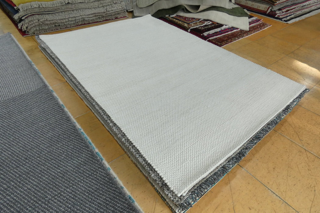 A large rectangular white rug with a textured woven surface and dark gray patterned edges, displayed on a light wood floor with other rugs visible in the background.