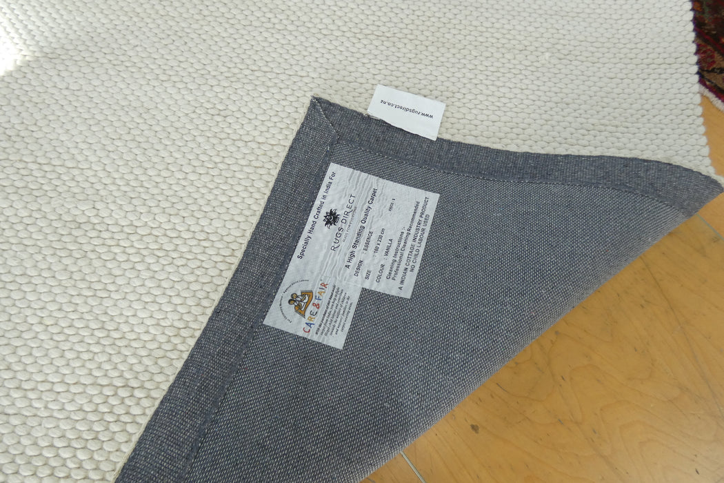 A gray woven area rug shown from above, folded over at one corner to reveal the underside and a brand label. The underside is heather gray, and the top surface is a textured cream/off-white.