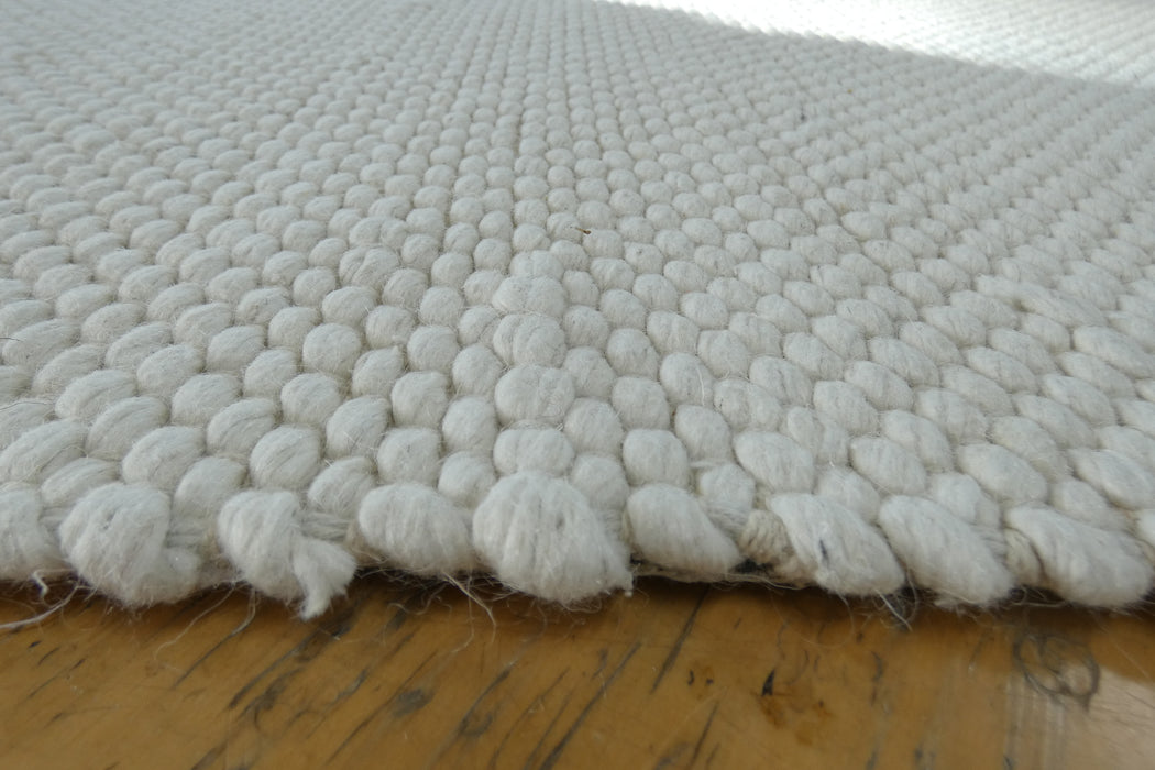 A close-up of a white textured area rug with a chunky, looped tufted pattern, shown resting on a wooden floor.