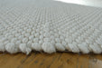 A close-up of a white textured area rug with a chunky, looped tufted pattern, shown resting on a wooden floor.