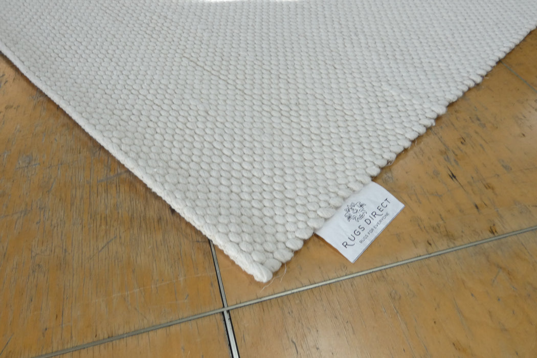 A close-up of a white textured area rug with a visible 'RUGS DIRECT' brand tag, shown on a light brown wooden floor.