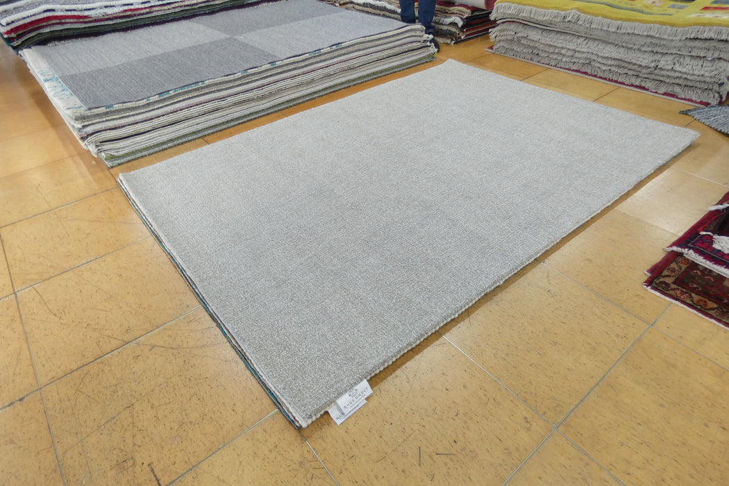 A light gray rectangular area rug displayed on a tan tiled floor, with stacks of other rugs visible in the background.