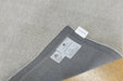 Close-up of a gray textured rug corner folded over to reveal the underside, with two white labels attached and a light wood floor visible underneath.