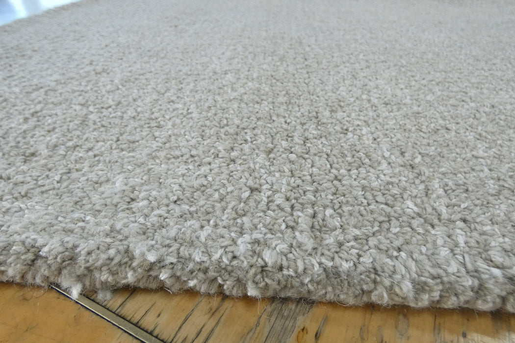 Close-up of a plush, high-pile shag rug in a light oatmeal gray color, showing the thick textured fibers and the underside with a thin metal binding edge resting on a wooden floor.