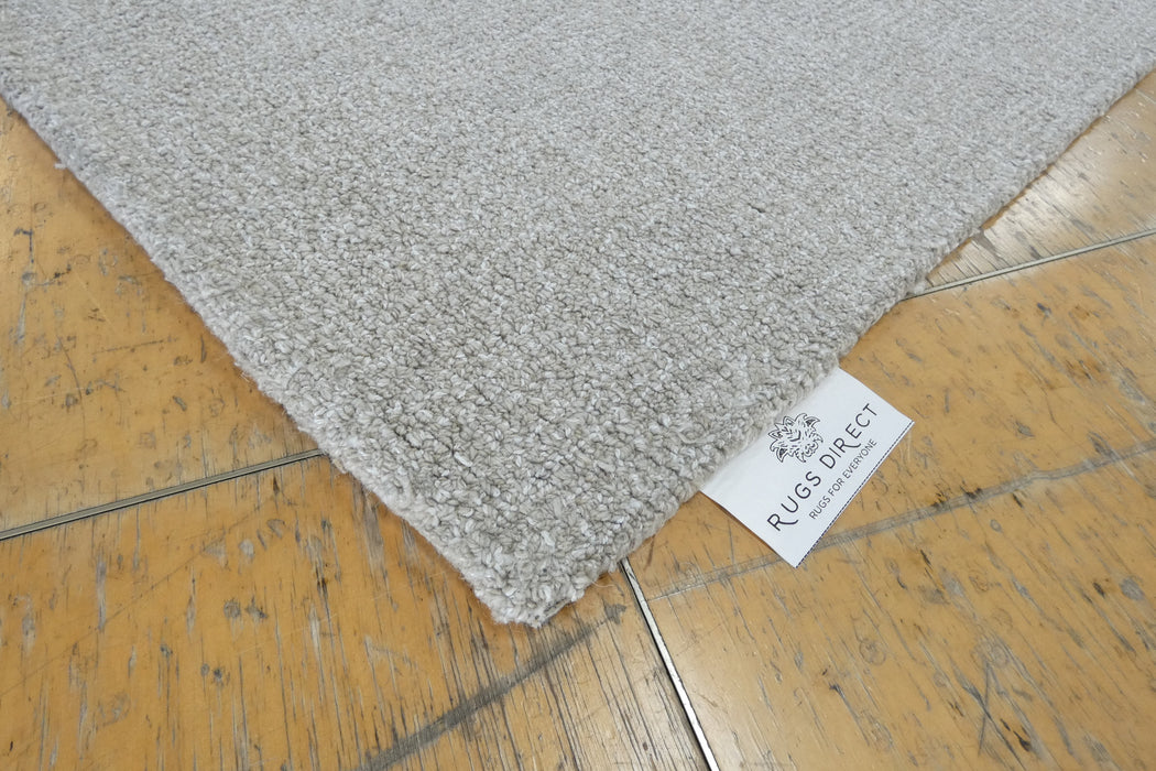 A close-up of a light gray textured rug corner placed on a yellow-brown wooden floor, with a white tag reading RUGS DIRECT visible at the edge.
