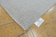 A close-up of a light gray textured rug corner placed on a yellow-brown wooden floor, with a white tag reading RUGS DIRECT visible at the edge.