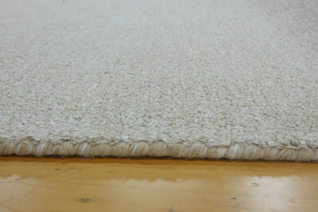 Close-up of a cream-colored shag rug with a thick textured pile and fringed edge, shown resting on a light wood floor.