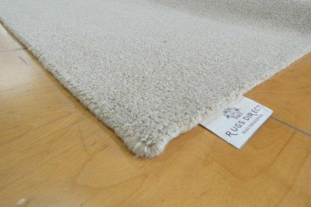 A close-up of a plush, high-pile area rug in a light cream or off-white color, laid out on a light wood floor. A white brand tag for 'RUGS DIRECT' is visible at the corner of the rug.