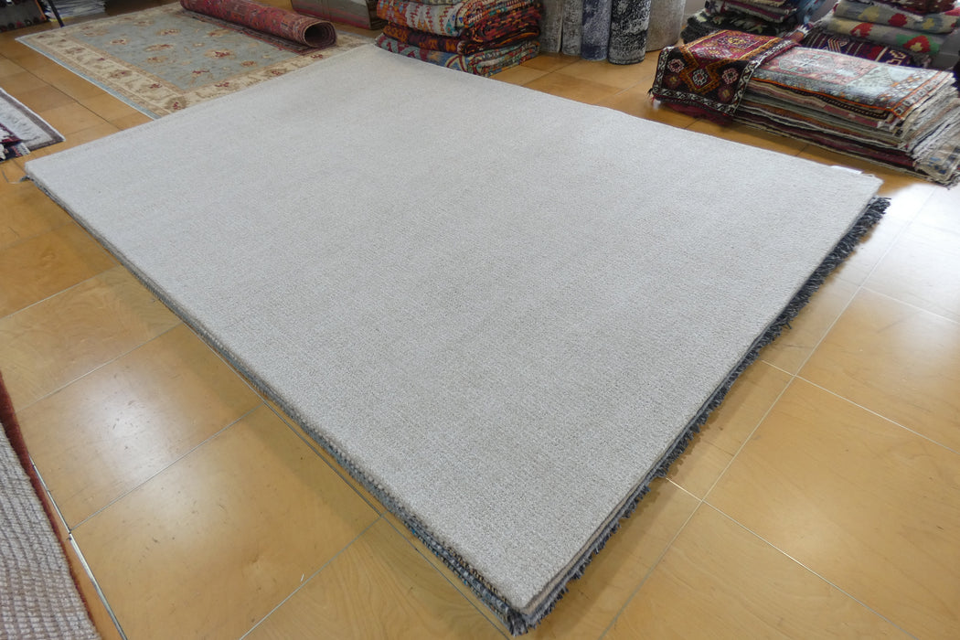 A large light gray rectangular rug or mat with a dark fringed edge, photographed on a polished wood floor in a showroom with other rugs and textiles visible in the background.