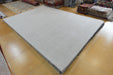 A large light gray rectangular rug or mat with a dark fringed edge, photographed on a polished wood floor in a showroom with other rugs and textiles visible in the background.