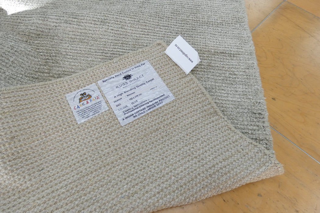 A close-up of a woven natural fiber rug or mat with two visible product labels, placed on a light wood floor. The label text includes 'Rug Factory' and '100% Natural Fibers'.