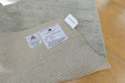 A close-up of a woven natural fiber rug or mat with two visible product labels, placed on a light wood floor. The label text includes 'Rug Factory' and '100% Natural Fibers'.