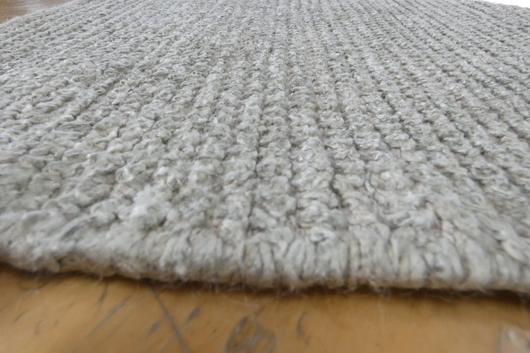 A close-up of a light gray textured rug with a chunky looped pile, shown folded over to reveal its thickness and underside, resting on a wooden floor.