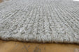 A close-up of a light gray textured rug with a chunky looped pile, shown folded over to reveal its thickness and underside, resting on a wooden floor.