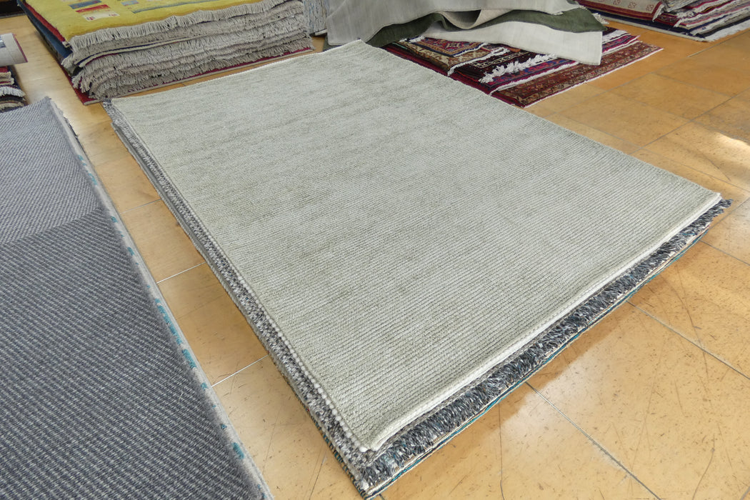A large rectangular area rug in a light gray and white ribbed pattern with a dark gray patterned border, displayed on a light wood floor in a showroom with other rugs visible.